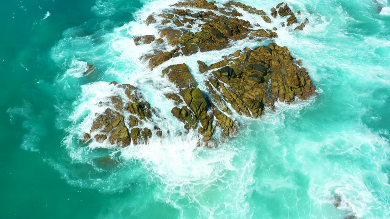 Overhead view of turquoise water and white foam wrapping around brown rock outcrops at Marquis Los Cabos, with no visible people.