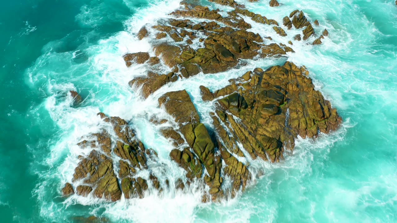 Top-down view of a larger brown rocky outcrop bordered by bright white foam and turquoise ocean at Marquis Los Cabos, with no visible people.