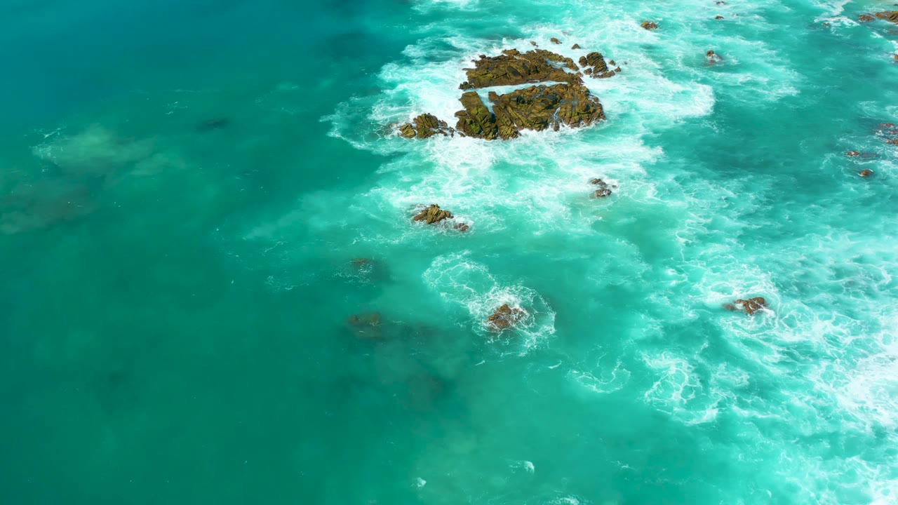 Aerial frame from Marquis Los Cabos with foamy surf spreading diagonally from the upper rocks into open turquoise water, with no visible people.