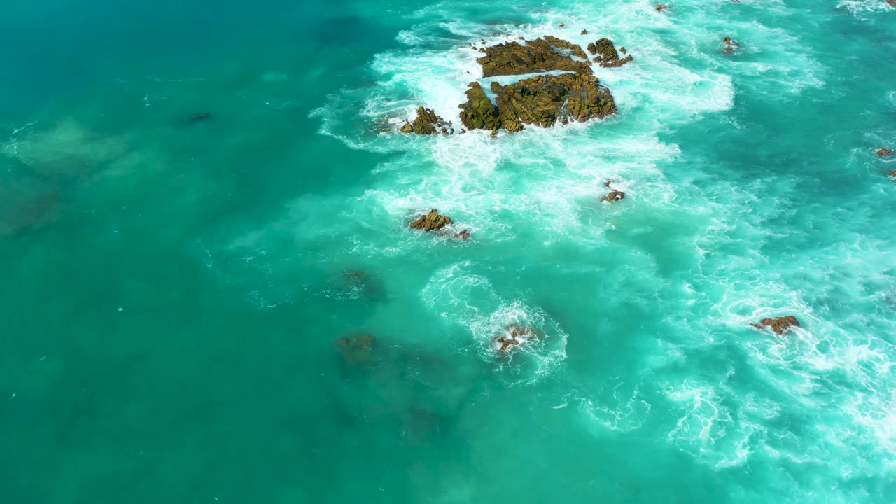 High drone view over Marquis Los Cabos water where a central rocky outcrop is ringed by white foam and smaller rocks sit in the surrounding turquoise sea, with no visible people.