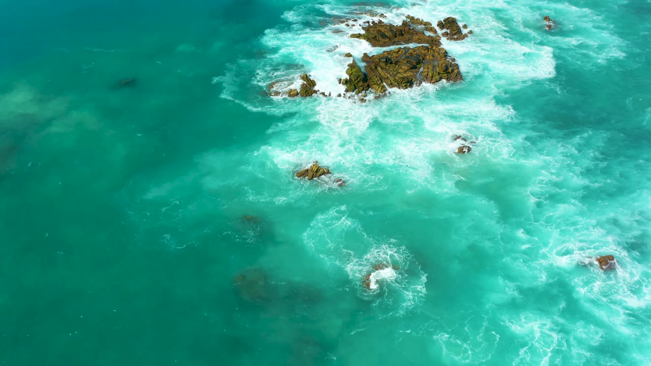 Marquis Los Cabos aerial frame with exposed brown rocks near the upper center and white surf curling around them across teal water, with no visible people.
