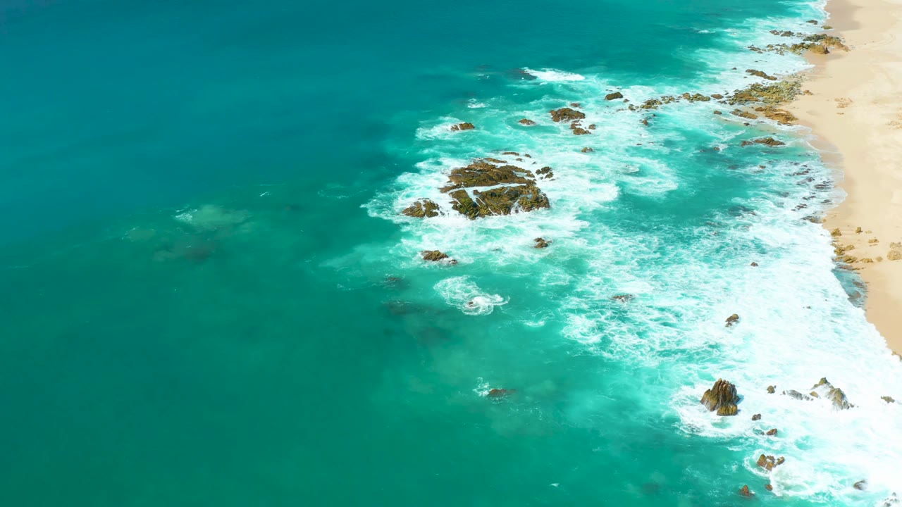 Aerial frame at Marquis Los Cabos, Mexico showing a larger white foam field around rocks, open teal water to the left, sand at the upper right, and no visible people.