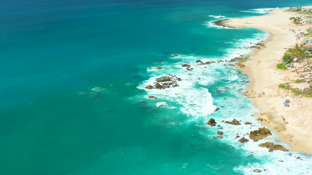 Aerial view at Marquis Los Cabos with a broad teal water field, foamy waves curling into the rocky shallows, beach running along the right edge, and no visible people.