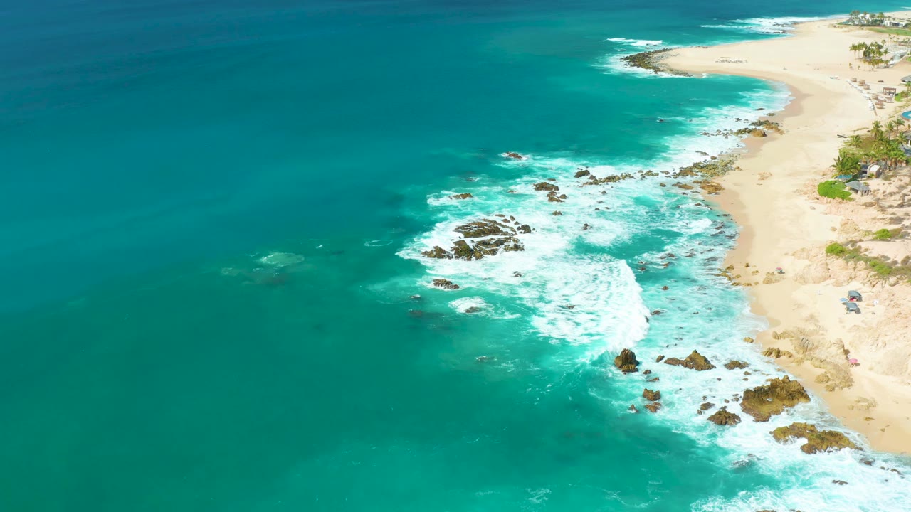Aerial frame over Marquis Los Cabos with turquoise ocean dominating the view, clustered rocks under white wash, a curved sandy beach at the right, and no visible people.