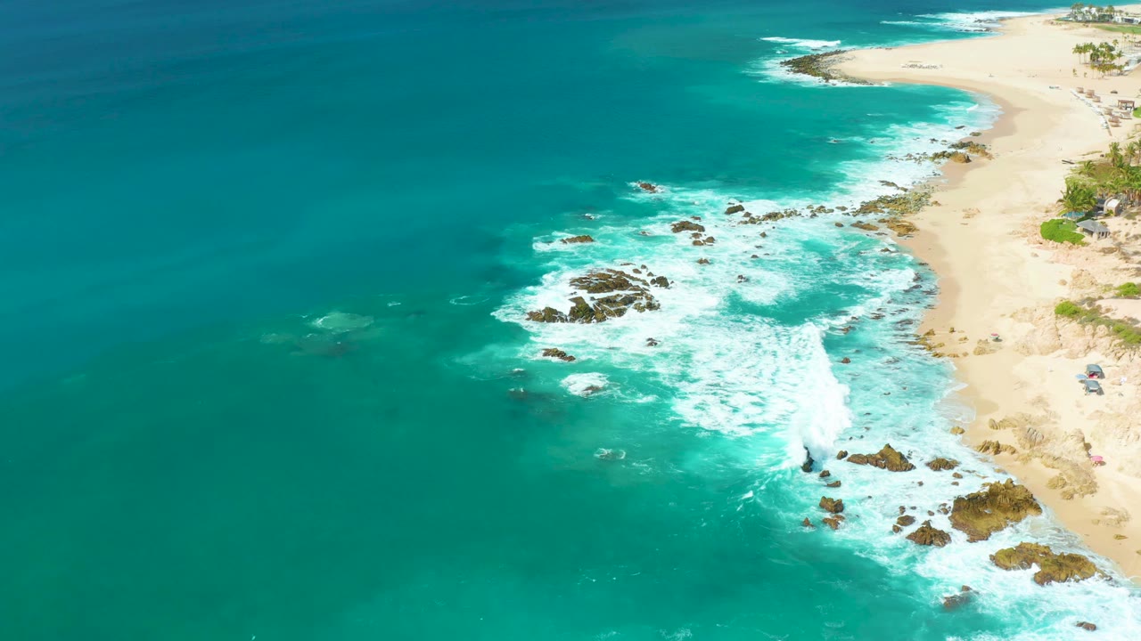 Mid-clip aerial view at Marquis Los Cabos where the foam has spread across the central rocks, with open turquoise water to the left, sand to the right, and no visible people.