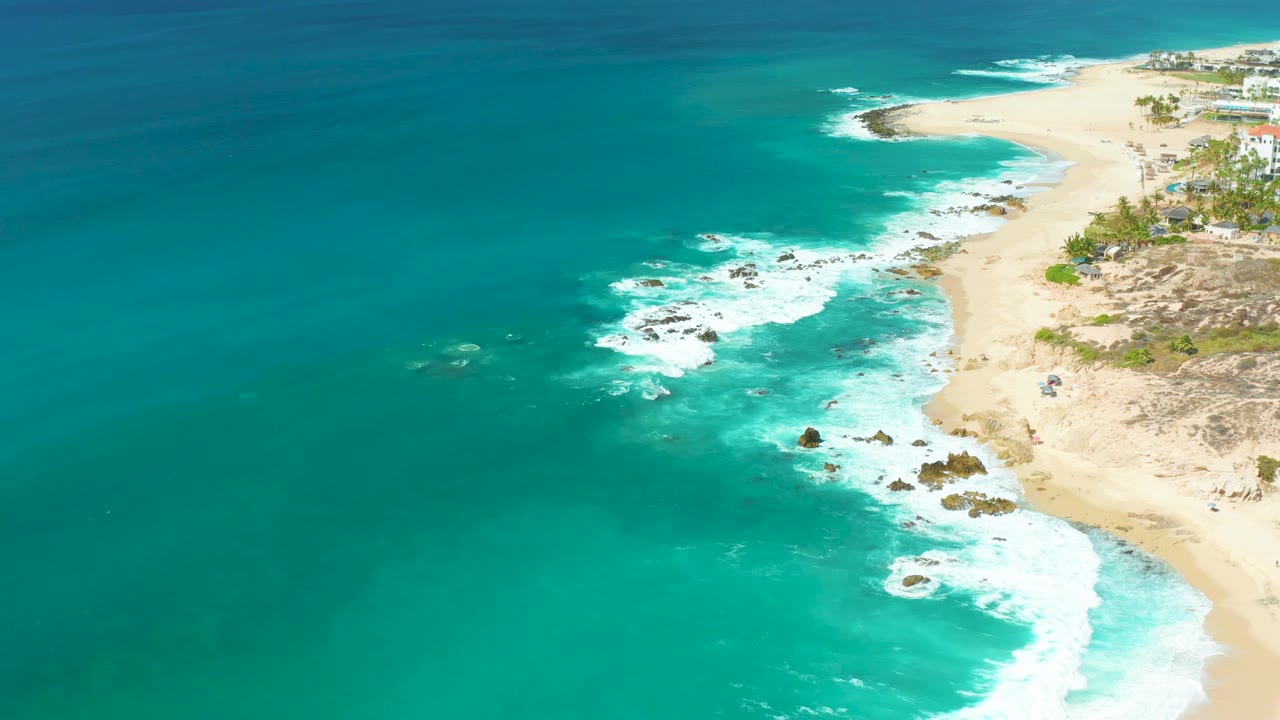 Aerial view at Marquis Los Cabos with surf spreading around dark rocks, pale sand curving up the right side, coastal buildings near the top right, and no visible people.