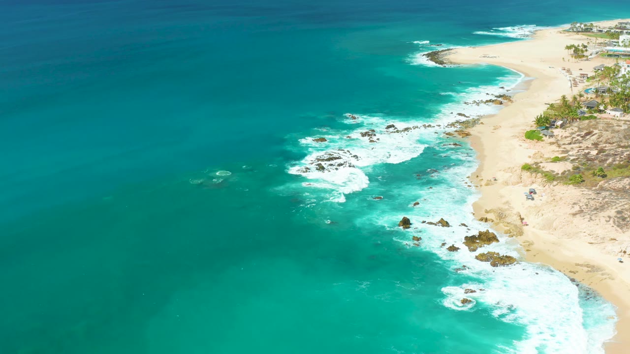 Wide aerial view of blue-green ocean beside a sandy beach with surf and rocks.