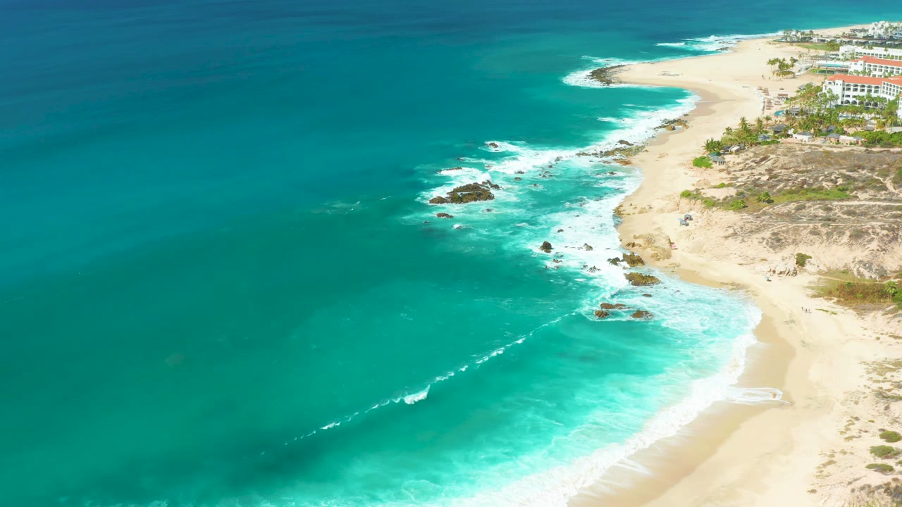At Marquis Los Cabos in Baja California Sur, turquoise water spreads across the left of the frame as white surf reaches rocks and pale sand, with no visible people.