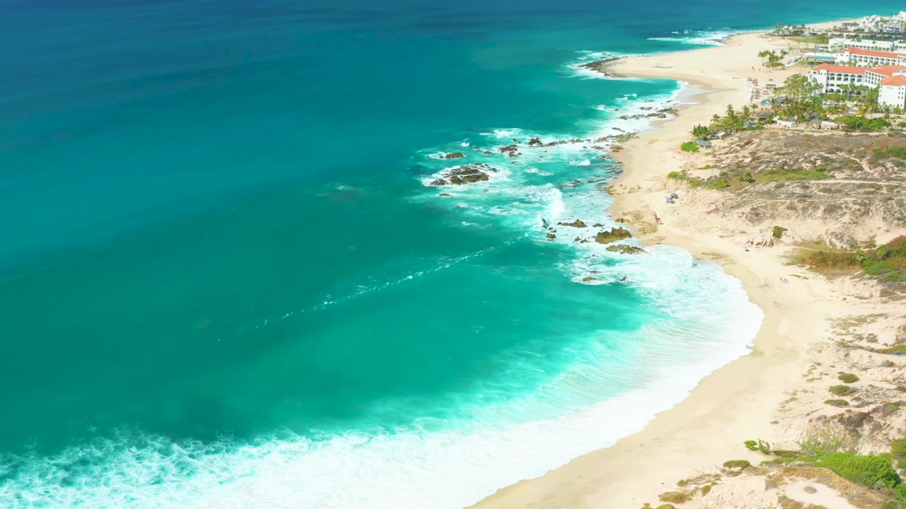 A second aerial frame holds on the Marquis Los Cabos shoreline as white foam spreads over the sand near dark rocks, with turquoise water offshore, distant buildings at upper right, and no visible people.
