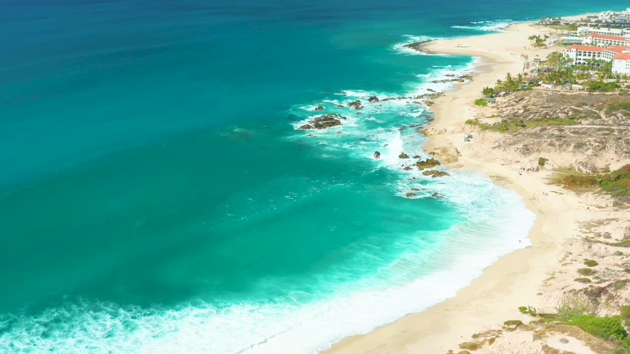 At mid-clip, white water breaks more visibly around the shoreline rocks while turquoise ocean fills the left side, the sandy beach runs upward into the distance, buildings sit near the top right, and no people are visible.