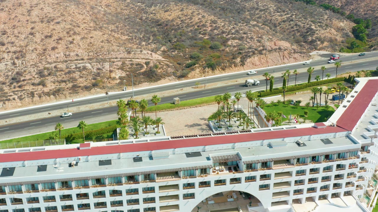 Aerial frame over Marquis Los Cabos showing a multi-lane highway, white resort building with a red roof edge, palm landscaping, dry hillside, moving cars, and no visible people.