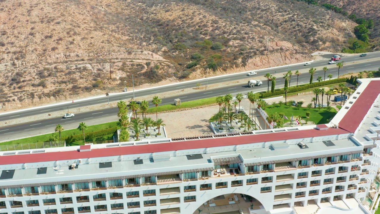 High aerial view at Marquis Los Cabos with cars moving along the highway, a landscaped palm area below, a white building roofline at the bottom, dry hills beyond, and no visible people.