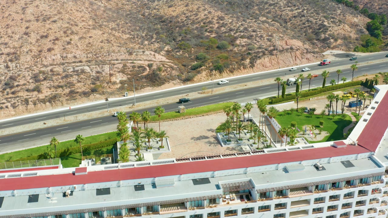 Aerial frame from Marquis Los Cabos showing the highway crossing the center, cars spaced along both directions, palm trees and green grass below, dry hillside above, and no visible people.