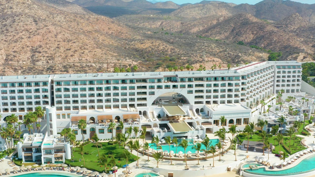 Aerial frame at Marquis Los Cabos with the white multi-story hotel centered, pool terraces and palms below, and rocky brown hills filling the background, with no visible people.
