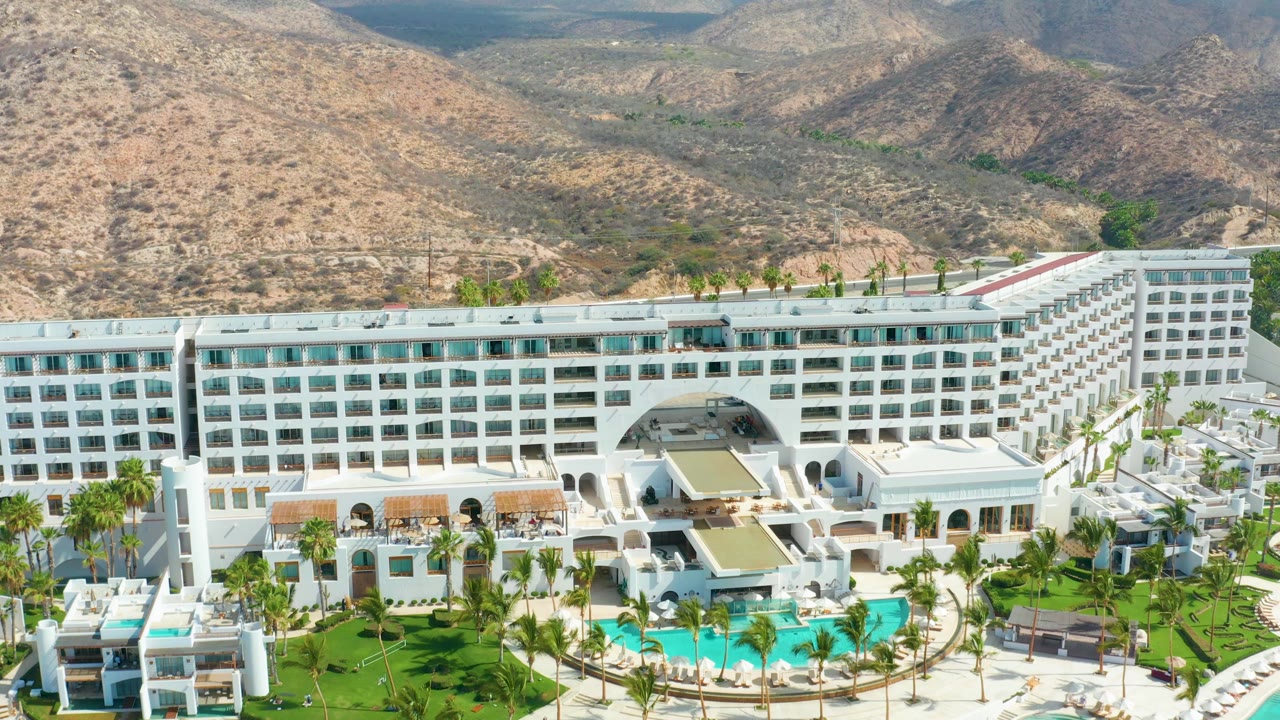 The drone view shifts along Marquis Los Cabos, showing more of the white hotel wing, landscaped lawns, pool water, palm rows, and arid mountain slopes, with no visible people.