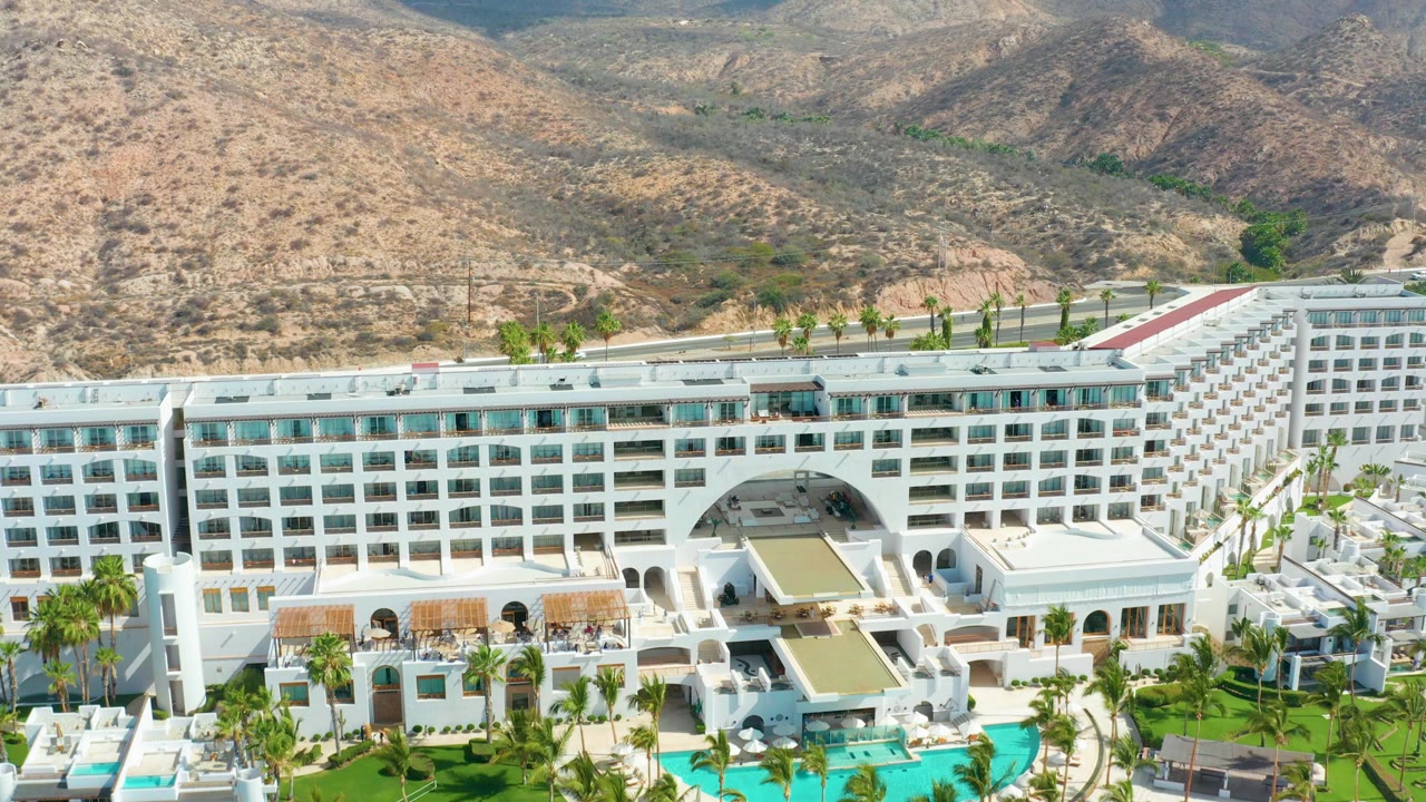 Mid-clip aerial at Marquis Los Cabos with the hotel roofline lower in frame, pools and palms near the foreground, a road behind the property, and dry hills beyond, with no visible people.