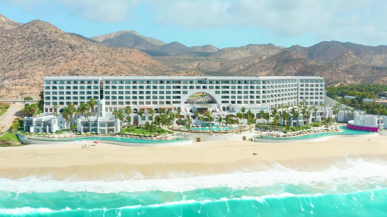 Opening frame of Marquis Los Cabos in Baja California Sur, showing a white beachfront hotel behind a pale sandy beach and turquoise surf, with resort pools, palms, arid mountains, blue sky, and no visible people.