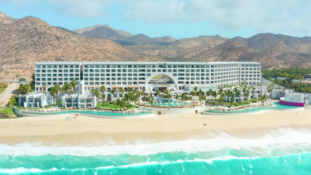 At 0:01, the Marquis Los Cabos beachfront remains centered while the turquoise waterline sits lower on the sand, with pools, palms, white hotel wings, arid mountains, and no visible people.