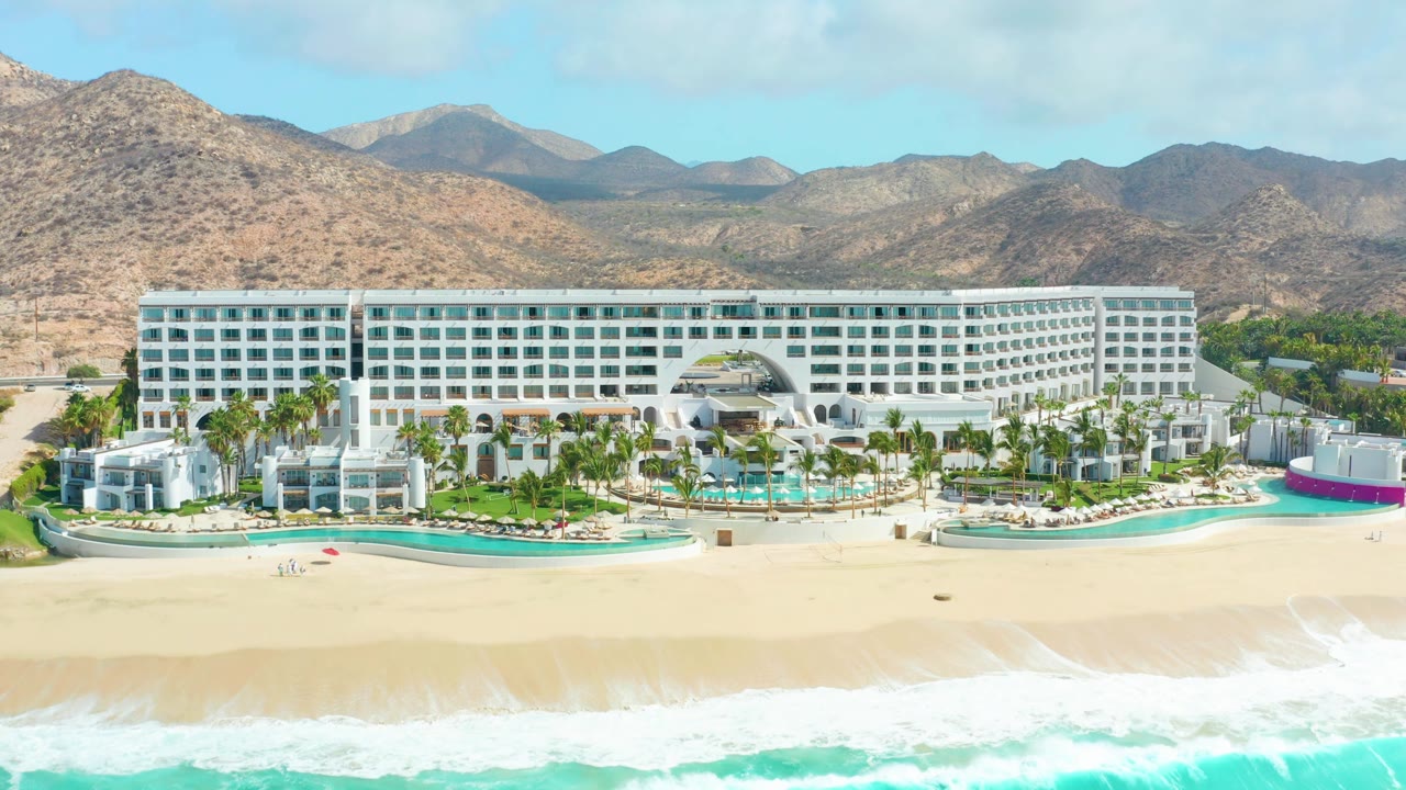 At 0:02, the wide aerial view holds on the Marquis Los Cabos hotel and beachfront, with the right-side hotel wing prominent, bright surf along the bottom edge, dry hills behind, and no visible people.