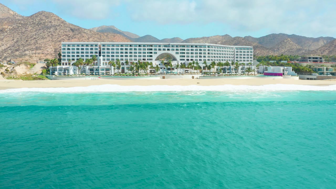 At 0:01, the aerial view holds on the Marquis Los Cabos hotel as the turquoise water covers most of the lower frame and a thin band of white surf reaches the sandy beach, with no visible people.