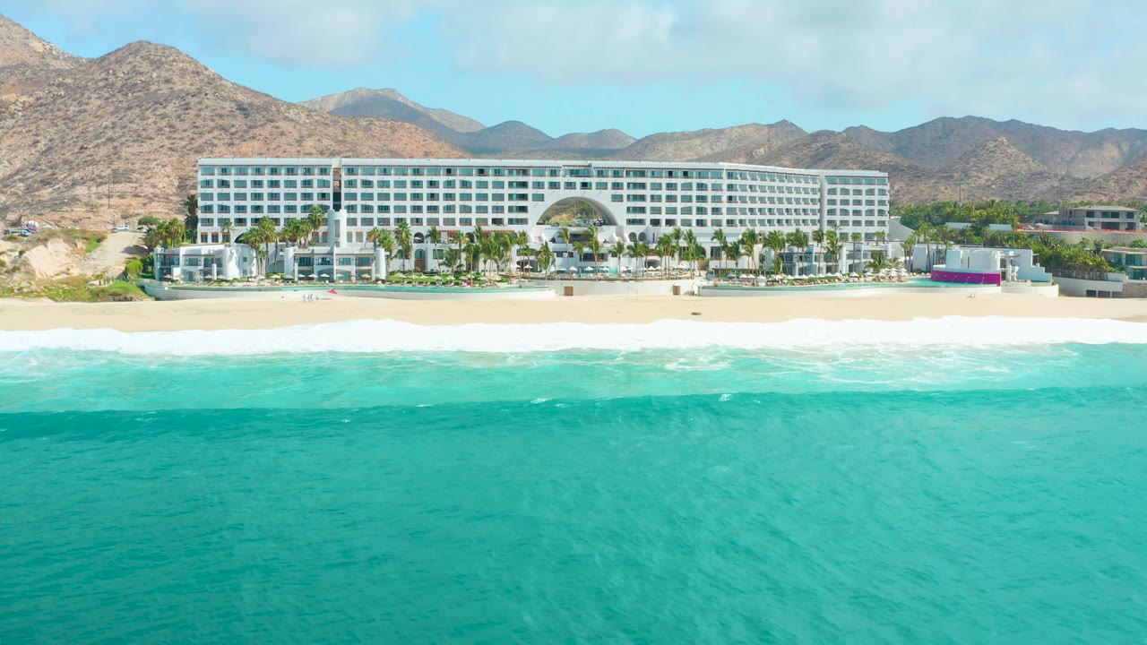 At 0:03, Marquis Los Cabos is seen closer across the shoreline as white foam spreads along the sand and turquoise water fills the foreground, with no visible people.
