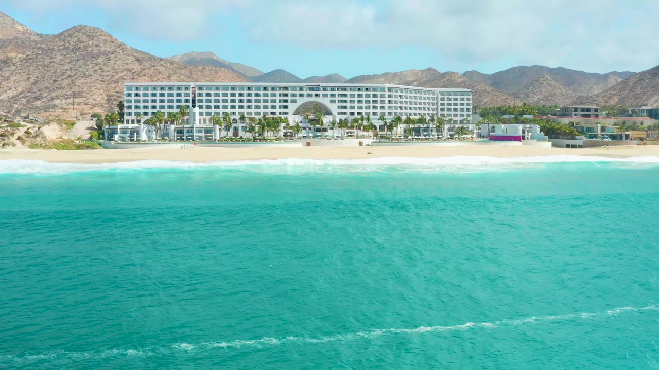 At 0:00, turquoise water fills the foreground below a sandy beach and large hotel at Marquis Los Cabos, with mountains and blue sky behind it and no visible people.