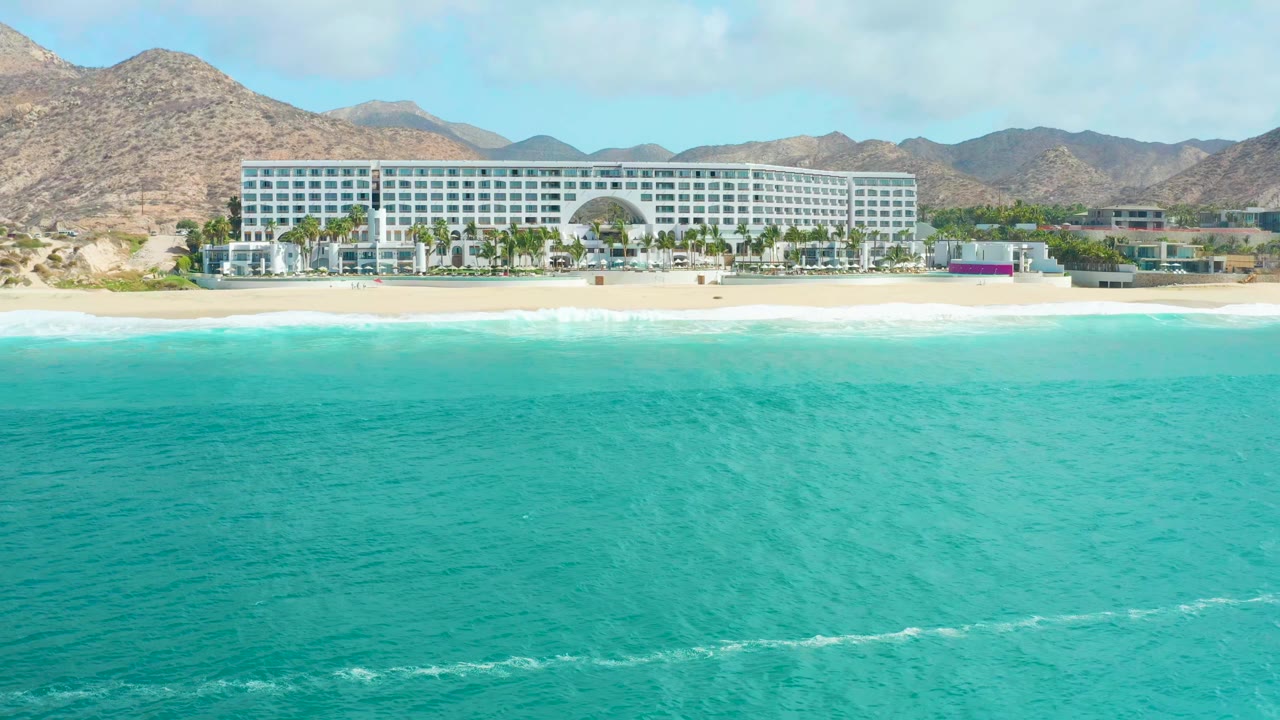 At 0:01, the wide view holds on turquoise ocean with faint wave lines, the sandy shore, the Marquis Los Cabos hotel, and brown mountains under a bright sky, with no visible people.