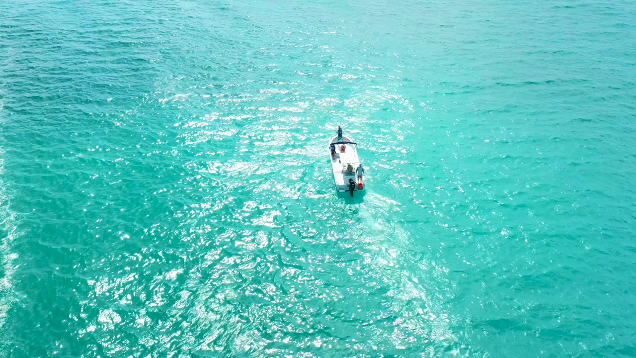 A high overhead view shows the small boat slightly farther through the turquoise sea, with a narrow wake and bright reflected light on the water surface.