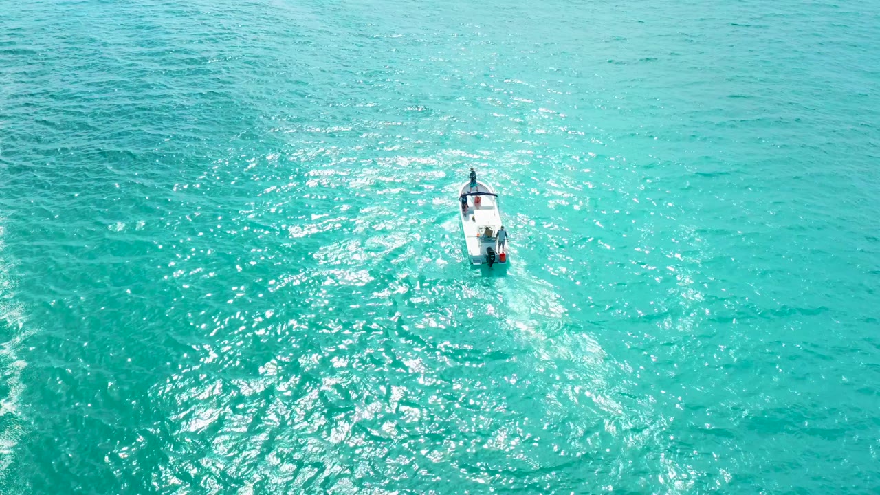 The boat remains small in the upper right portion of the aerial frame, moving through textured turquoise water with a pale wake behind it.
