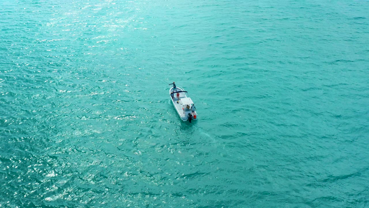 A small boat appears farther across the overhead frame on turquoise water in Marquis Los Cabos, with darker ripple patches below it and no visible people.