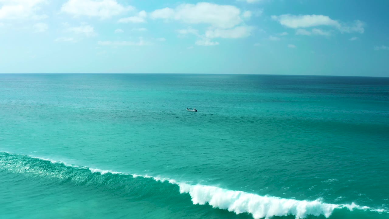 Wide elevated view of turquoise ocean at Marquis Los Cabos, Mexico, with a white wave cresting across the lower frame, a small distant boat near the horizon, and no visible people.