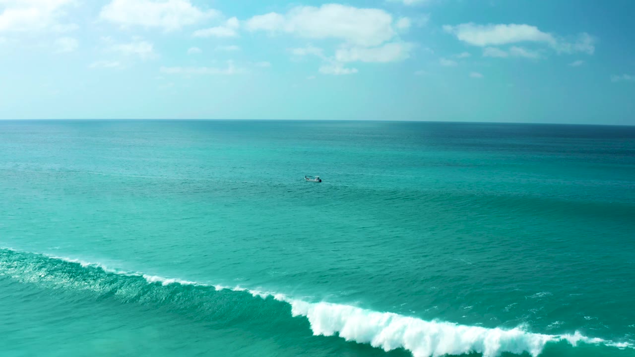 Wide ocean frame from Marquis Los Cabos with white foam spreading along the lower edge, clear turquoise water through the midground, a small boat offshore, and no visible people.