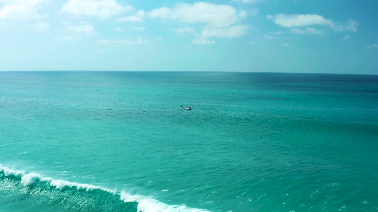 At Marquis Los Cabos, the breaking wave sits lower in the frame with foam thinning, broad turquoise water beyond it, a small distant boat, pale sky, and no visible people.