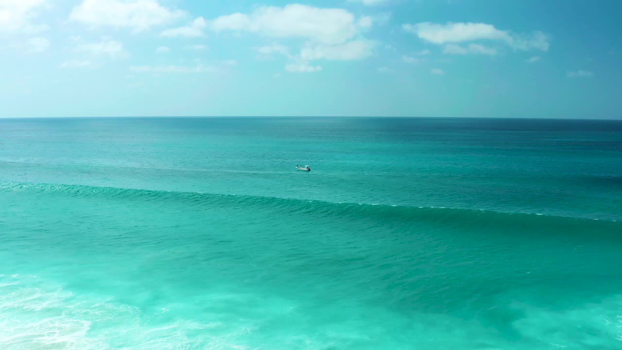 The Marquis Los Cabos ocean view holds a high horizon, layered blue-green water, a continuous forming wave in the lower frame, a tiny distant craft, and no visible people.