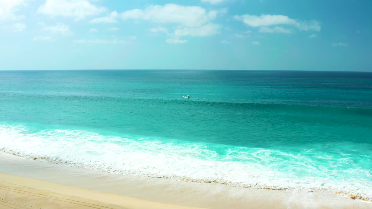 Aerial view at Marquis Los Cabos, Mexico, with turquoise water, white foam, pale sand at the bottom edge, a level horizon, scattered clouds, and no recognizable people visible.