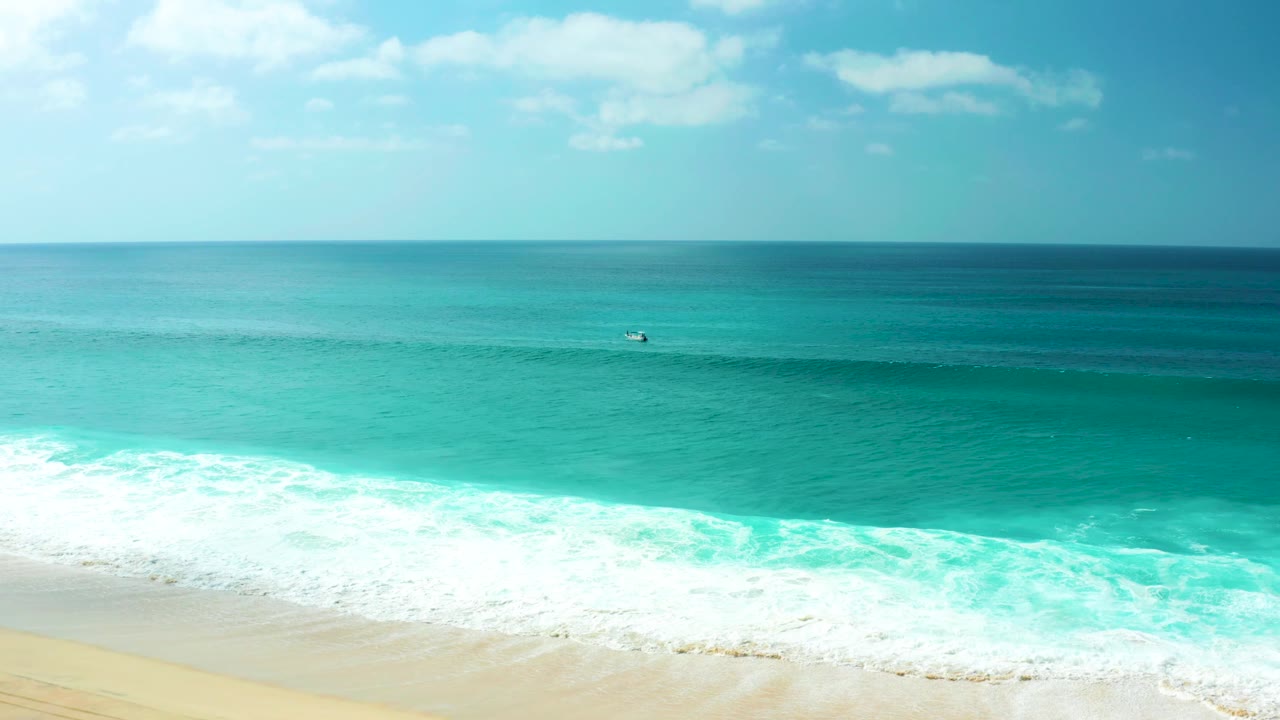 Marquis Los Cabos ocean view with a broad band of turquoise water, active white foam at the lower left, pale sand below, and a tiny distant surfer or board too small to identify.