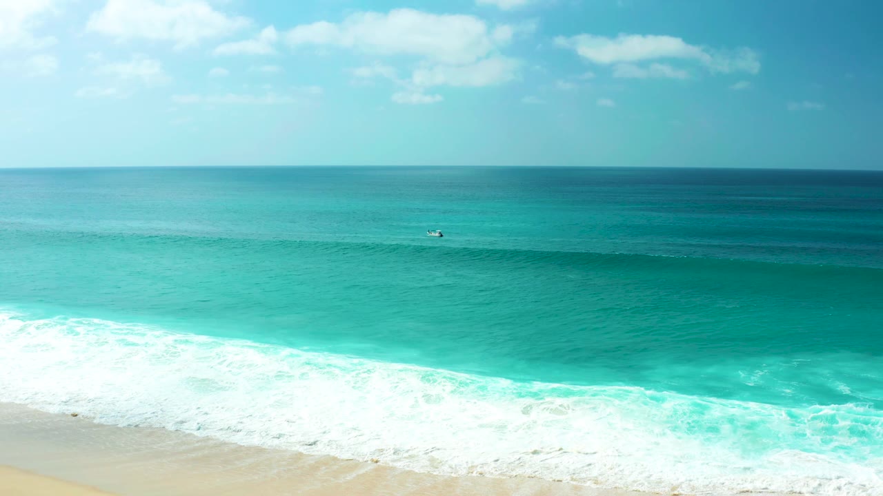 Aerial beach view with turquoise water, white foam, and a small dark surfer offshore.