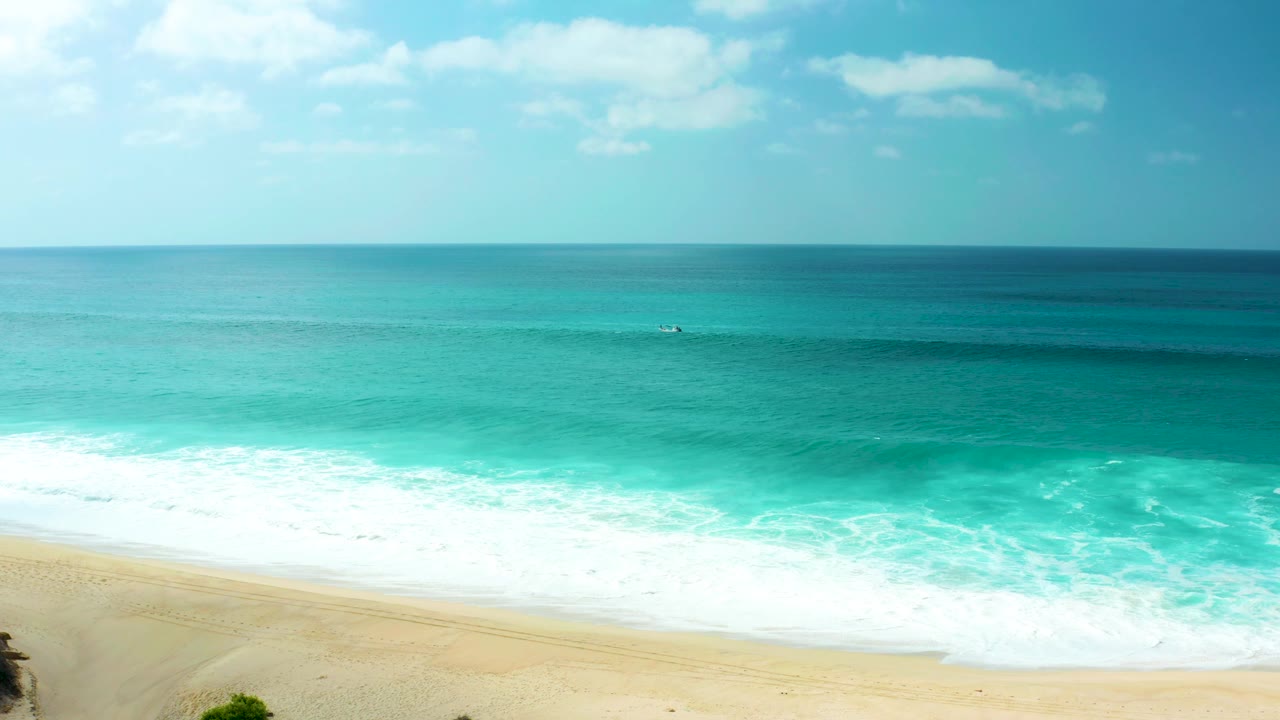 An elevated view shows blue-green water moving toward the empty Marquis Los Cabos sand, with a soft cloud patch above the level horizon and no visible people.