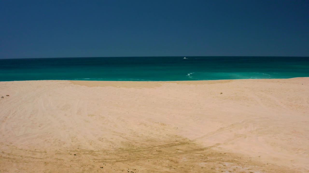 Empty sandy shoreline at Dreams Los Cabos with turquoise water near shore, deeper blue water offshore, a straight horizon, and no visible people.