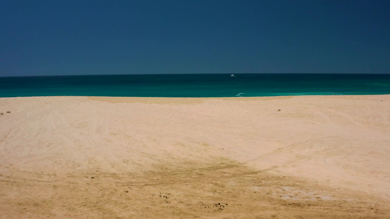 Broad beach foreground at Dreams Los Cabos with faint sand patches, a thin turquoise surf zone, darker offshore water, clear sky, and no visible people.