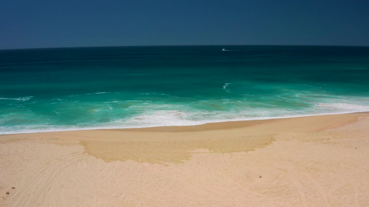 A low band of white foam spreads along the Dreams Los Cabos shoreline, with pale sand below, teal water above, and no visible people.