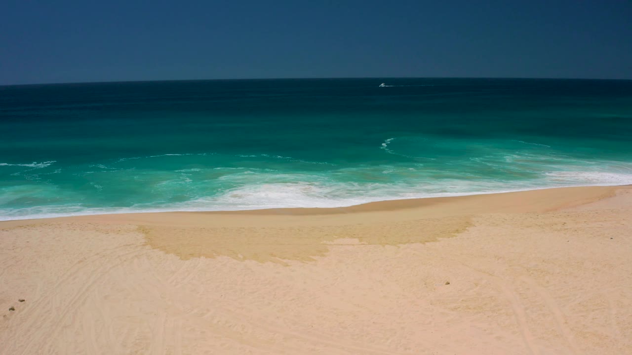 The wave has receded slightly at Dreams Los Cabos, leaving a wider patch of wet sand between the dry beach and turquoise water, with no visible people.