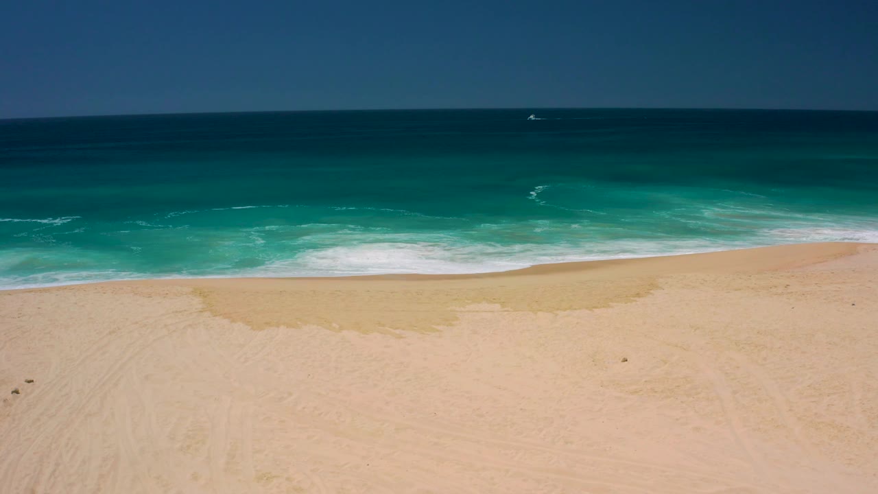 An empty stretch of Dreams Los Cabos beach fills the lower frame while turquoise surf rolls across the middle and the horizon stays level, with no visible people.