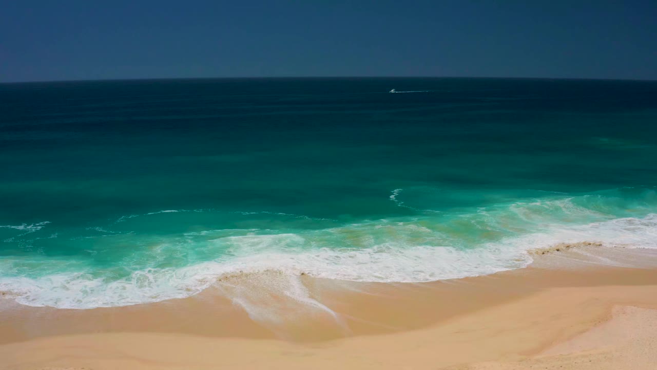 At Dreams Los Cabos, white foam spreads farther across the center of the frame while turquoise water and empty sand remain visible with no visible people.