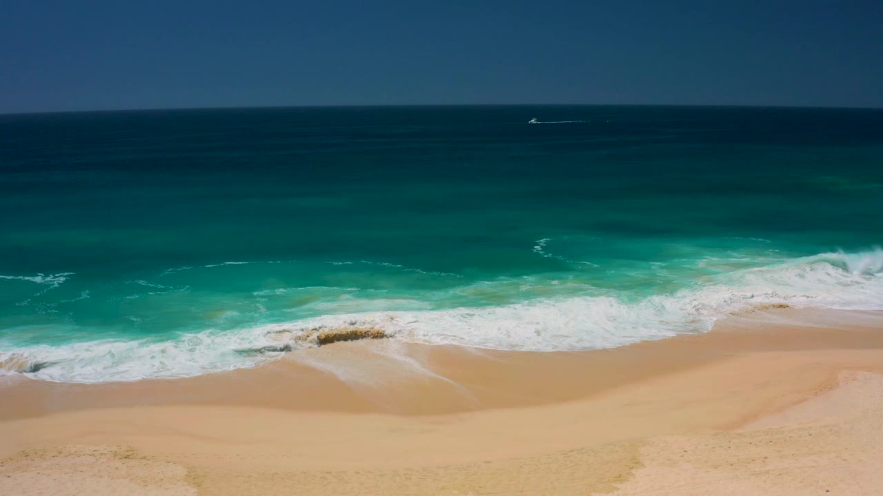 The Dreams Los Cabos beach appears from above as foam curls onto pale sand, with deep blue water beyond the surf and no visible people in frame.