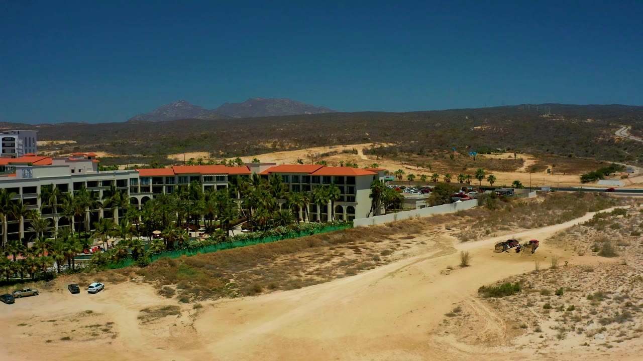 Aerial frame at Dreams Los Cabos with more of the palm-lined building centered, parked cars near the grounds, dry scrubland and dirt path in front, mountains on the horizon, and no visible people.