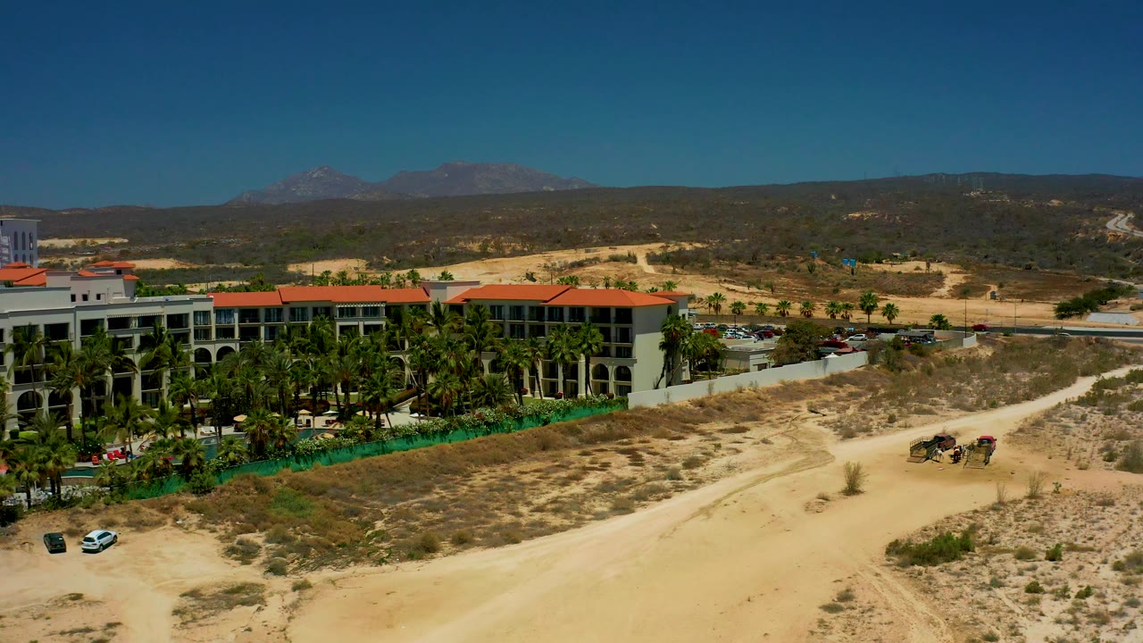 Mid-clip aerial frame at Dreams Los Cabos with the building shifted farther left, palm trees and green fencing below it, a white car near the dirt track, mountains beyond, and no visible people.
