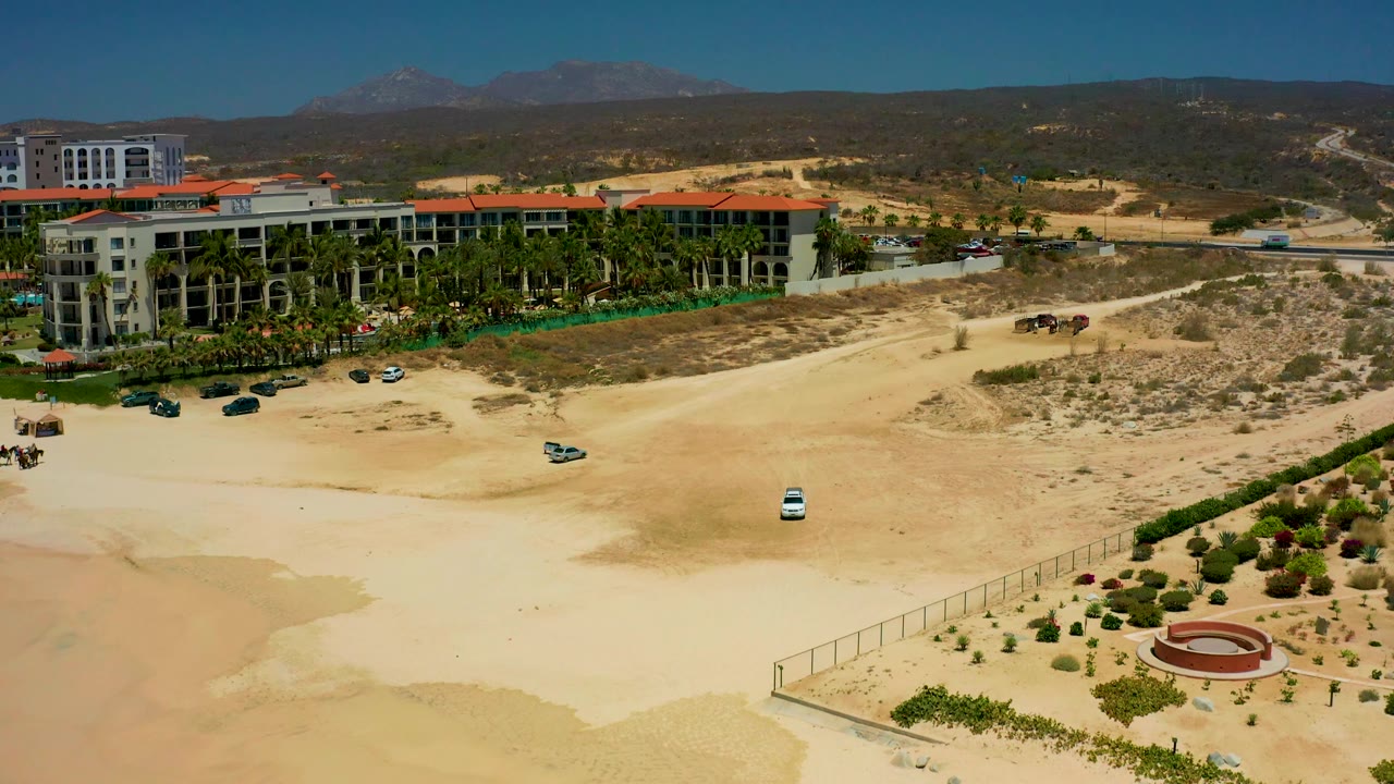 Aerial view at Dreams Los Cabos with the white car farther along the sandy road, resort buildings to the left, a fenced planted area below, mountains beyond, and no visible people.
