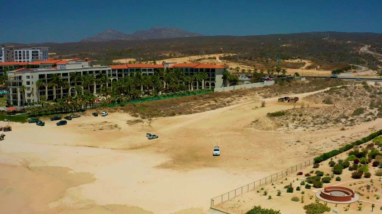Wide Dreams Los Cabos aerial frame with red-roof buildings sliding toward the left side, sandy roads and dry scrub across the foreground, the white car on open ground, and no visible people.
