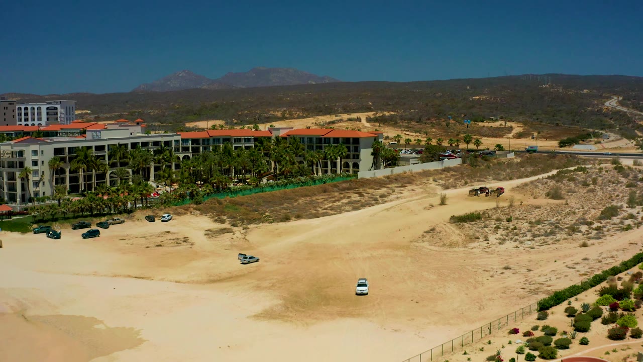 Mid-clip aerial frame at Dreams Los Cabos showing the red-roof resort block along the left edge, sandy access roads and dry brush across the center, mountains on the horizon, and no visible people.